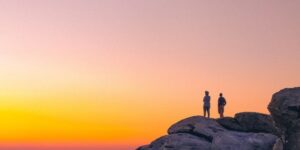 Two people standing on a large rock watching the sunset