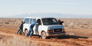 Three people pushing a broken down van on a dirt road