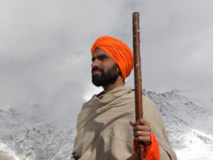 A man wearing an orange turban holding a staff with mountains in the background