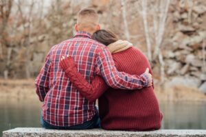 A man and woman sitting together on a rock staring at a pond with the arms around eachother