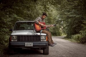 A man sitting on a car playing the acoustic guitar
