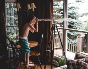  A young boy standing on a chair looking out of a window through a telescope