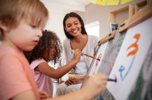 two young children painting on easels with a woman smiling at their work