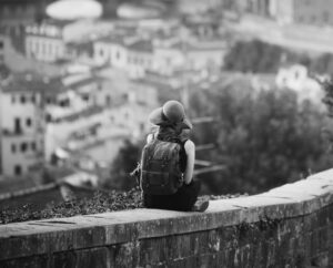 woman with a backpack wearing a hat sitting on a wall looking at a city